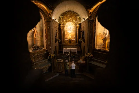 Two visitors facing an illuminated altar inside an ornate, warmly lit chapel.