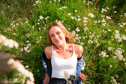 A woman sitting among white wildflowers and tall grass, looking up toward the camera.