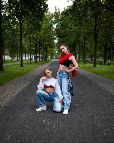 Two women posing on a tree-lined path, one crouching and one standing in blue jeans.